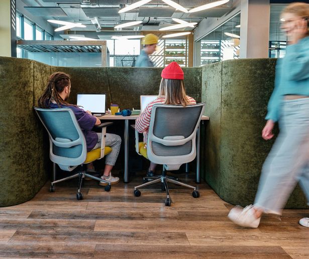 The post shows a busy office scene where people are rushing around on the phone. In the background there are two individuals sat looking at their laptops facing away from the camera. They are busy at work and sitting in tall office chairs with wheels.