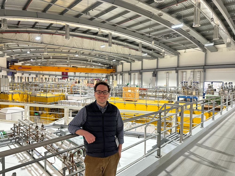 I’m standing on a walkway in the Diamond Light Source facility. The hall I am standing in is in the shape of a ring, and a portion of it — with exposed ducts and pipes — is visible behind me. A yellow beamline station can be seen behind me, on a level below the one I am on.