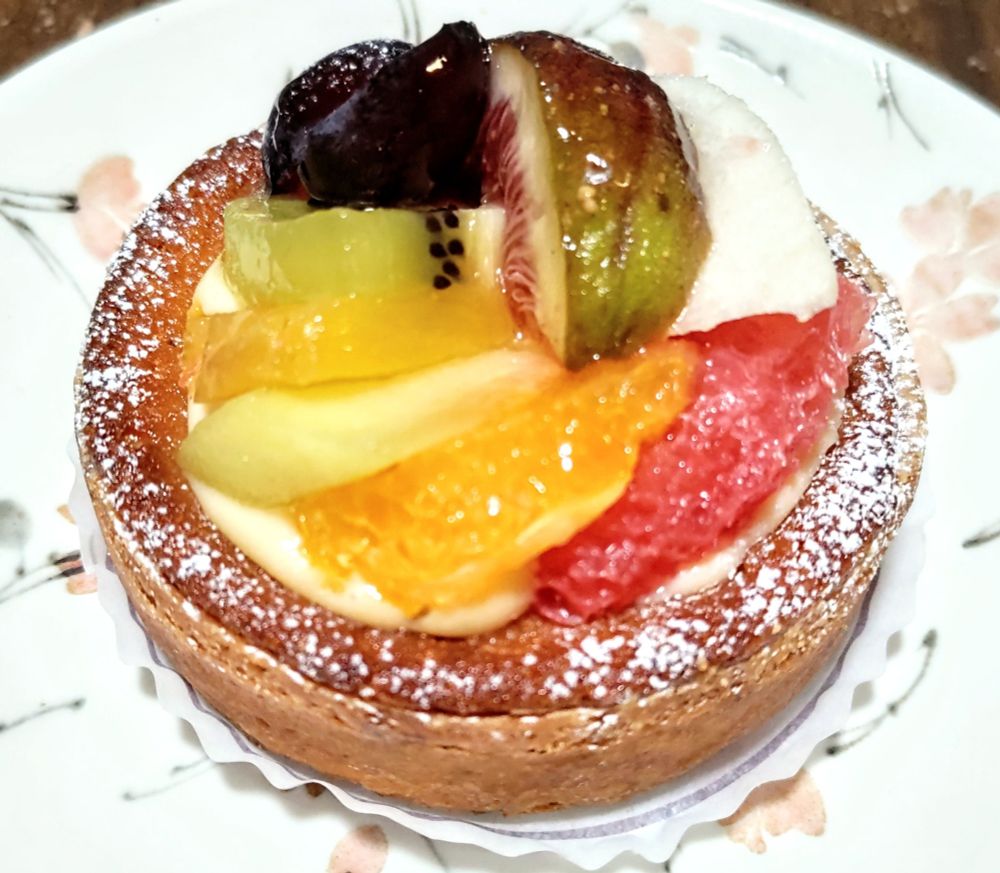 フルーツタルトの写真。
桜の花びら柄の皿の上にフルーツタルトが乗っている。
土台のタルトの上にはグレープフルーツ、オレンジ、メロン、柿、キウイ、ブドウ、イチジクが円形に並べられている。