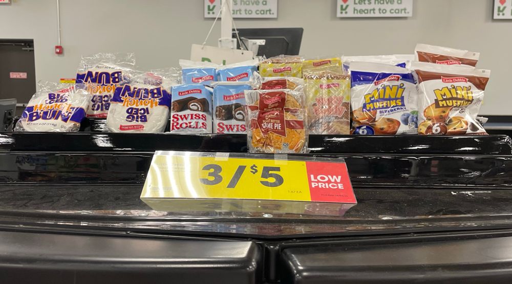 Sign in grocery store that reads: “3/$5” with Little Debbie snacks behind it.