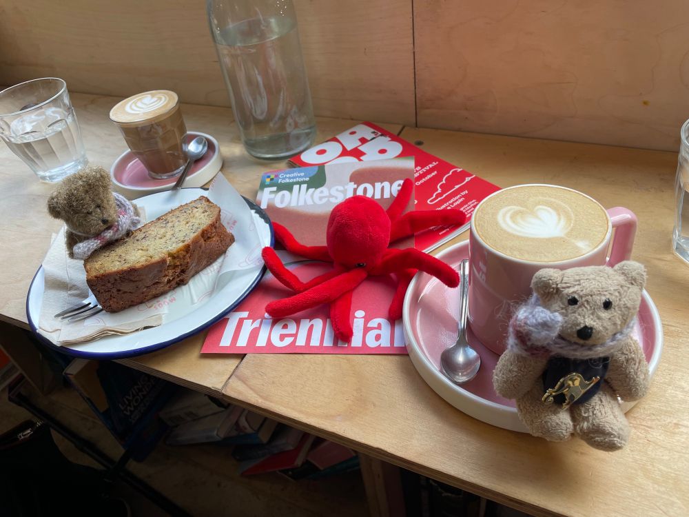 A cafe counter with cake and pink coffee cups, two plush bears and an octopus are on the counter with a flyer for the Folkestone Triennale arts festival 