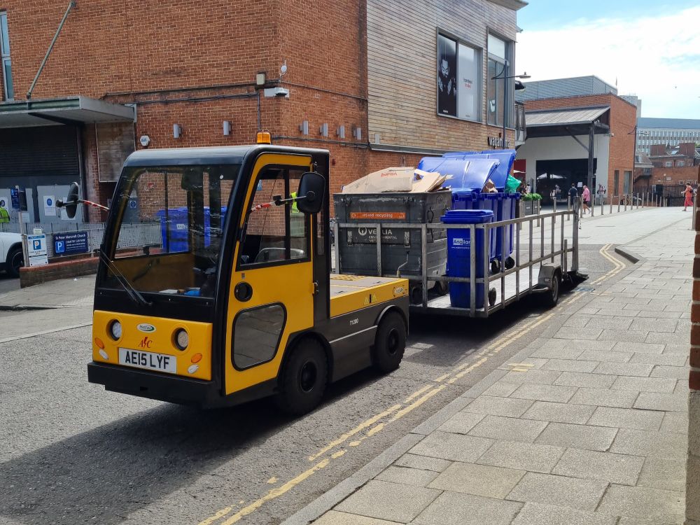 A small yellow and black electric 'locomotive' pulls a large trailer, on which the full paladins and wheelie bins are loaded. Presumably empty ones were taken off first to replace them. 
