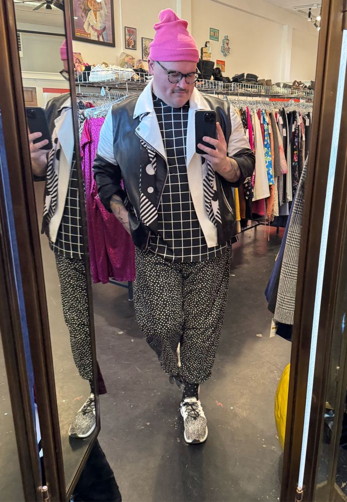 A homosexual takes a selfie in a mirror wearing a mix of clashing black and white patterns and a pink hat.