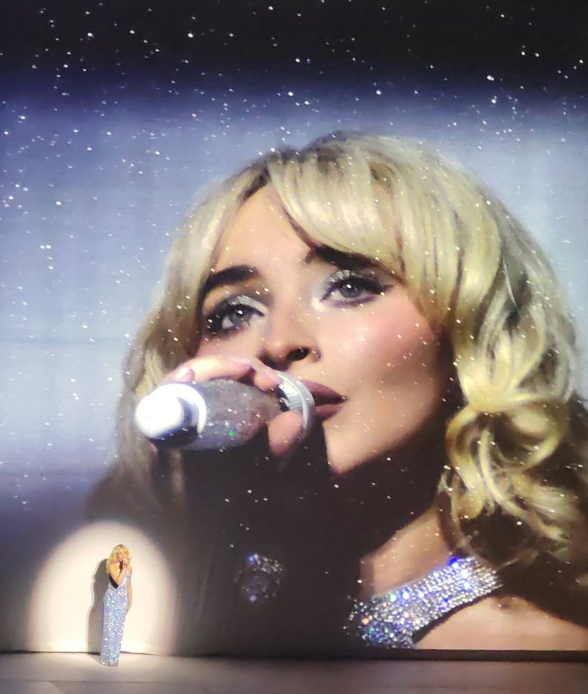 A photo of Sabrina Carpenter singing on stage in a bejeweled silver floor-length dress with a spotlight on her, and a larger close-up of Sabrina projected on the wall behind her. The closer look shows that her microphone and eye makeup are also similarly bejeweled in silver!