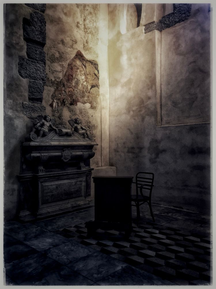 in the hushed solitude of the duomo di napoli, time lingers in quiet reverence. a single wooden desk and an empty chair rest upon the cold, checkered marble floor; silent witnesses to the passage of centuries, to prayers whispered and long forgotten. the air is thick with the weight of memory, the stone walls stained with history.

to the left, a grand tomb stands adorned with cherubic figures, their frozen expressions caught between sorrow and serenity. they guard the slumber of the departed, their marble forms resisting the erosion of time. above them, remnants of a faded fresco cling to the wall like a distant echo of divine artistry, its colors muted, as though the past itself is dissolving into the present.

light filters in through an unseen source, soft yet piercing, illuminating the rough textures of the ancient masonry. shadows stretch and contract, playing a silent symphony of light and darkness, presence and absence, the eternal dance of mortality.

this space, at once sacred and abandoned, invites contemplation. it is a chamber of paradox; a resting place that speaks, a room empty yet full, a fragment of eternity where the living and the dead converse without words. here, one feels the weight of impermanence, the poetry of decay, and the quiet promise that, in the grand stillness of time, all things return to dust, yet never truly disappear.