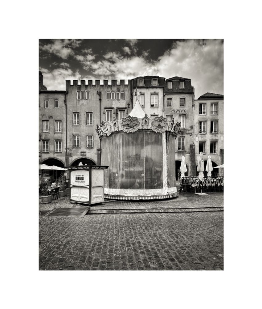 a carousel wrapped in veils, waiting in a deserted square.
its music silenced, its horses unseen. behind it, old facades watch like quiet witnesses, their windows the eyes of another century. cobblestones stretch forward, heavy with the memory of countless steps, yet no foot disturbs their silence now.

this is a portrait of potential, suspended: joy enclosed, time on pause. even the sky seems unsure.
clouds gathering, dispersing, unable to decide whether to break or to hold.

here, the carousel becomes a metaphor for longing: the longing for motion, for laughter, for the ordinary magic of turning in circles under an indifferent sky. we glimpse the fragility of human rituals, how easily they fold into stillness, and how in that stillness, they quietly endure, waiting to be called back to life.