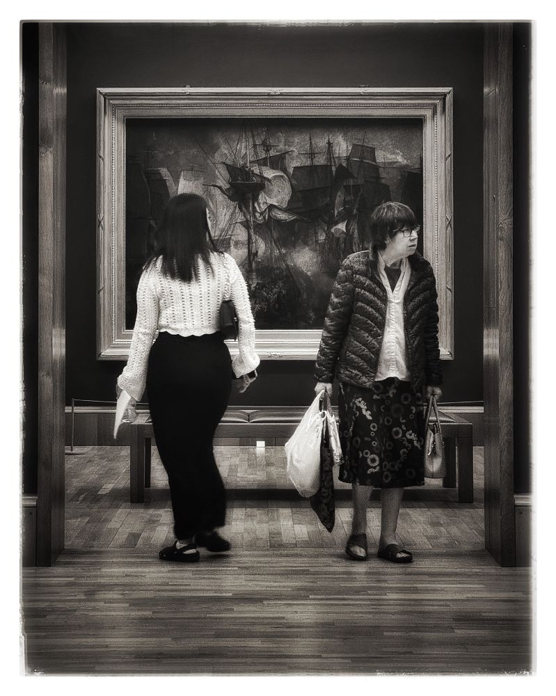in the hush of tate britain,
two human figures drift like opposing tides.
one drawn into the room of turner’s storm of sail and smoke,
the other anchored in the weight of plastic and days lived.

a ghost fleet swells behind them,
ships dissolving into sea-light and memory,
brushstrokes unraveling history
as if time were oil on canvas, fluid, fading.

their backs to each other,
they mirror disconnection,
yet hold a silent symmetry
in gesture, in pause,
in being caught 
between past and passing.

the floor beneath them is still,
but the painting moves.
and in that subtle motion,
turner speaks through shadow and salt,
as if to remind:
we are all only passing through the frame.