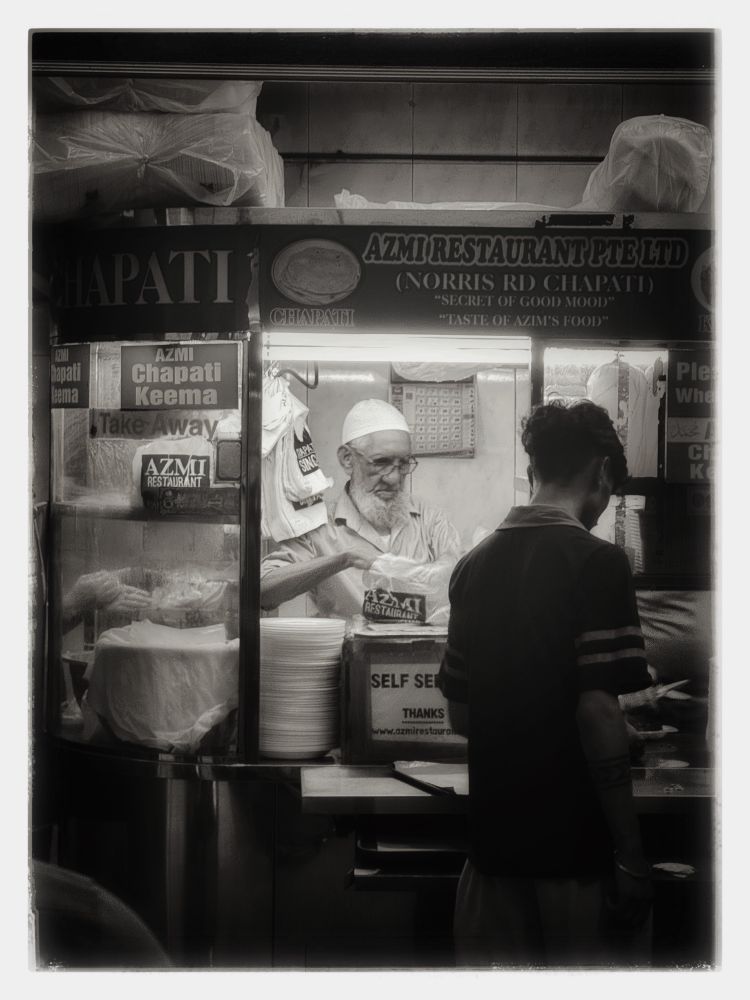 in this image, time stands still; not in the sense that nothing changes, but in the way a rhythm, once found, continues without haste. the scene, framed in black and white, renders a modest corner of little india into something sacred: a continuity of gesture, of labor, of quiet presence.

the man behind the counter, with his white cap and weathered hands, becomes more than a vendor, he is a custodian of memory. his actions are not merely economic but ritualistic, shaped by repetition and care. the chapati is not just food; it is the vessel of belonging, of story, of ancestral warmth folded into dough.

what’s remarkable is not the nostalgia, but the persistence. in a city that constantly reinvents itself, this place remains unchanged, not by resistance, but by fidelity to something older than progress. perhaps it is fidelity to slowness, to touch, to the trust between strangers that forms over hot meals and worn counters.

and so, returning here after fifteen years, you do not find the past, but a continuity, like a phrase that was never interrupted, merely paused until you were ready to listen again.