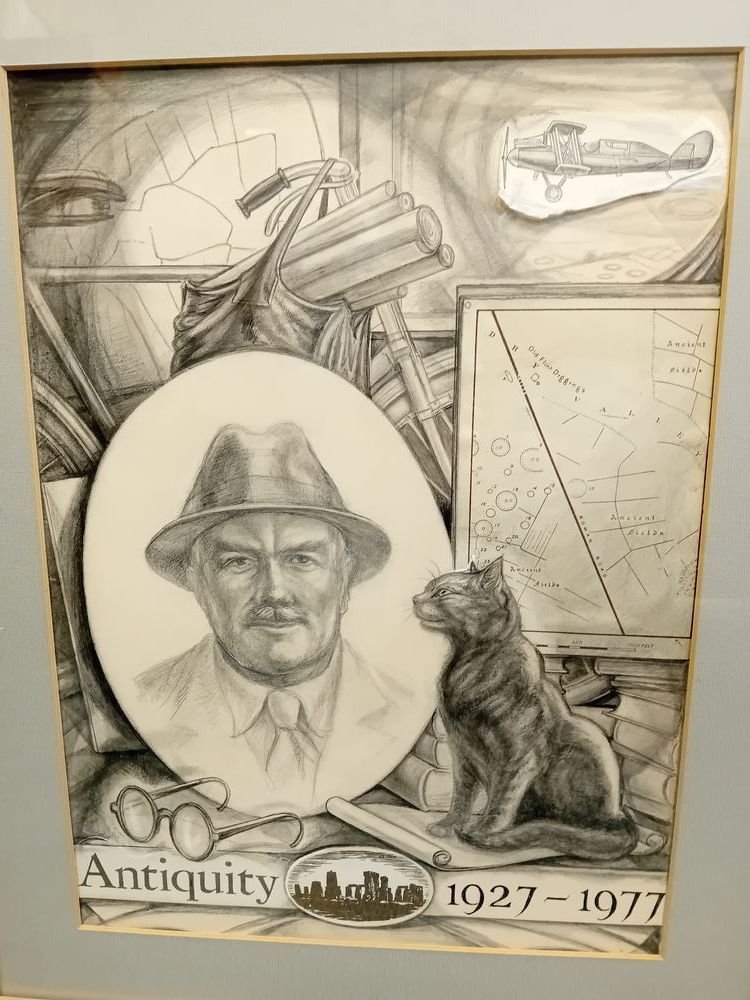 Illustration of a desk covered in papers, with a mirror reflecting a man in a suit and hat. A cat sits next to the mirror, alongside a map of earthworks and a plane with a propellor. Text at the bottom reads 'Antiquity 1927-1977'.
