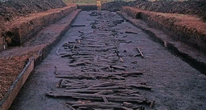 Rectangular trench in a muddy environment, in which sit many wooden planks and logs in situ, arranged to form a trackway.