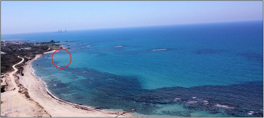 Aerial photograph of a bay. A sandy beach borders a clear blue sea. A red circle indicates the location of the shipwreck. 