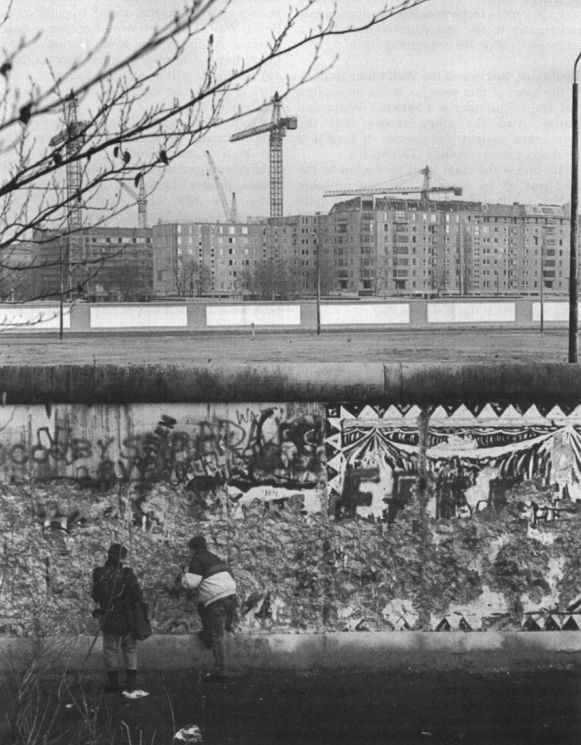 Black-and-white photograph of 'wall-peckers': two people chipping pieces from a graffitied section of the Berlin Wall to sell to tourists. Under-construction blocks of flats surrounded by cranes stand in the background (credit: Gerlind Klemens, 1990).