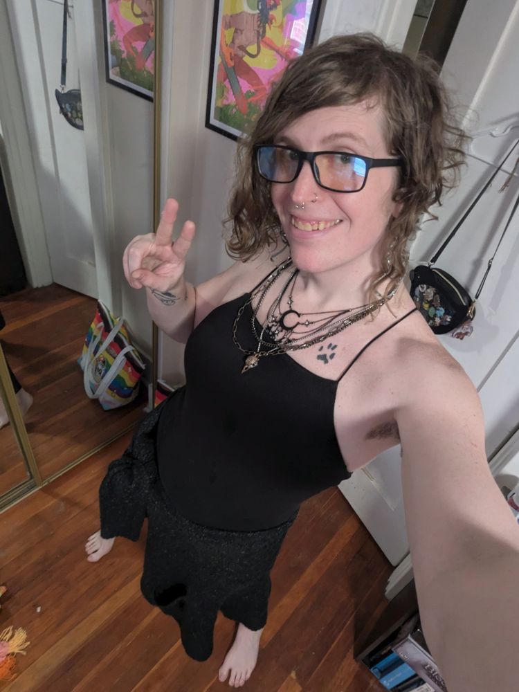 Selfie of Robin wearing a black dress, lots of necklaces, and a cardigan tied around their waist