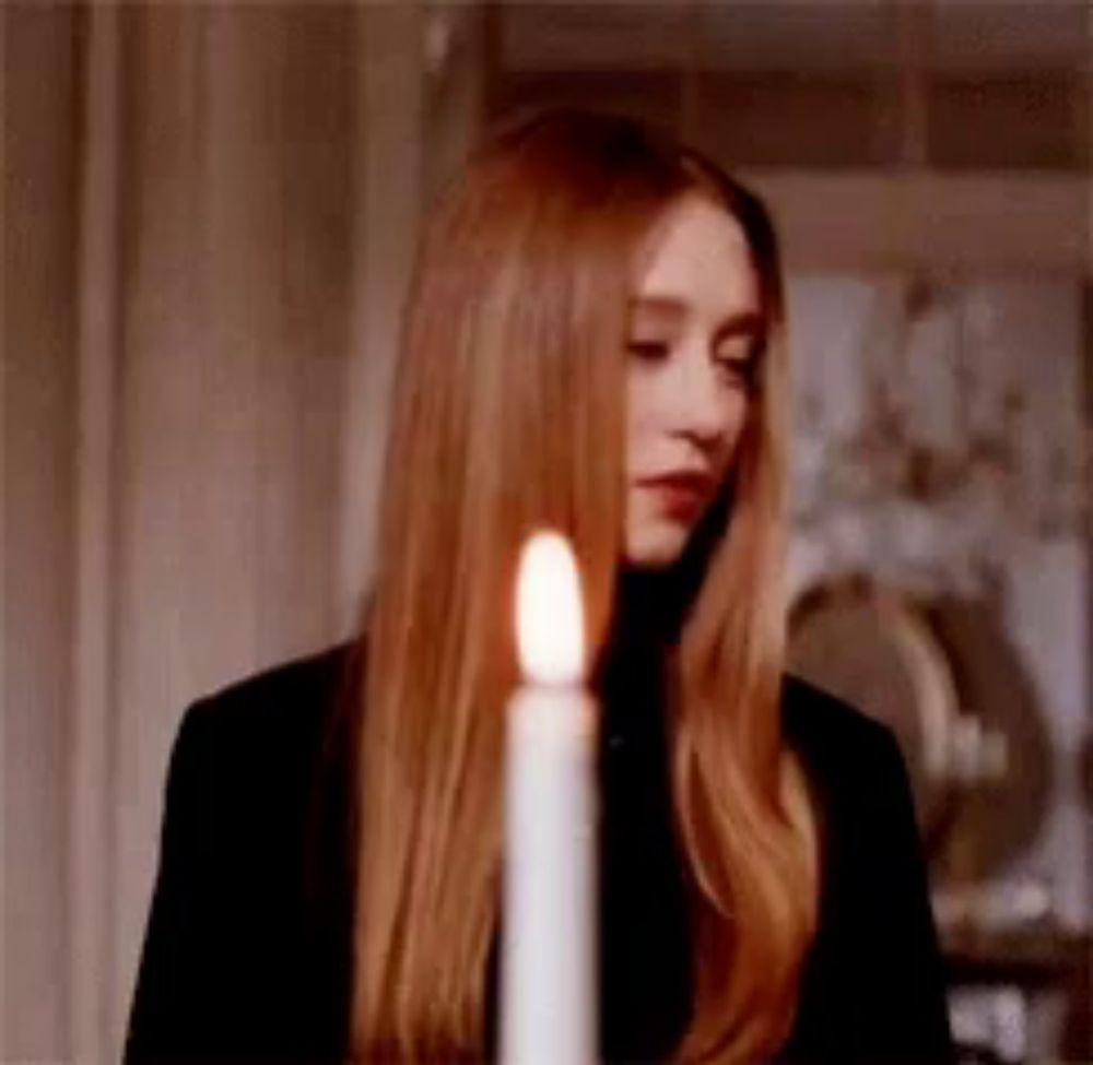 a woman with long red hair is standing in front of a lit candle