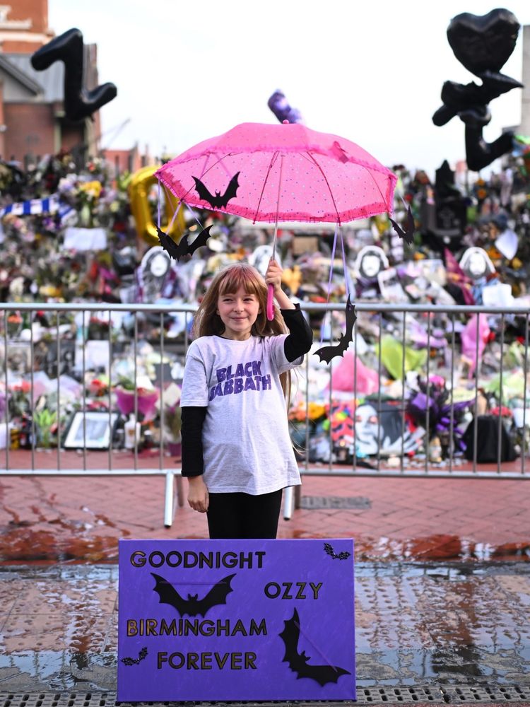 Child in Black Sabbath T Shirt holding a pink umbrella.  Purple sign with black bats on it says Goodnight Ozzy Birmingham Forever
 