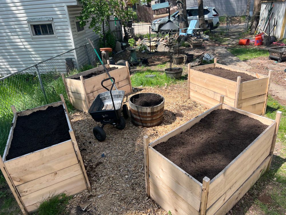 Four raised beds in a diamond configuration with a half whiskey barrel in the middle are filled with dirt ready for planting. A black utility wagon is visible