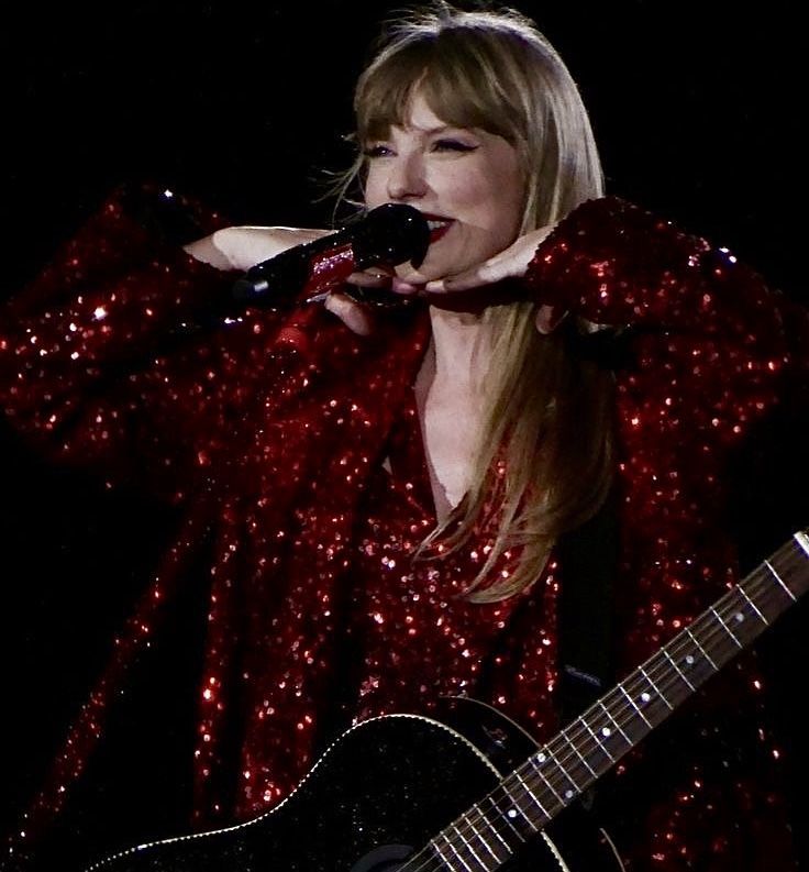 Taylor Swift smiling. She’s wearing a red glitter outfit with a black guitar. I think she’s performing at her tour 