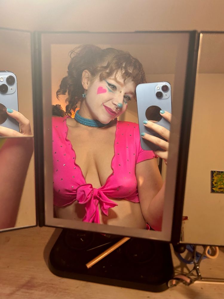 Woman with curly hair in a ponytail is taking a mirror selfie. she is wearing clown makeup that has big pink hearts for blush, and a blue nose. She is wearing a blue choker and a pink v-neck croptop. She has a septum piercing and a bridge piercing 

skyhub altgirl egirl clown clowngirl clown makeup pierced girl piercings 