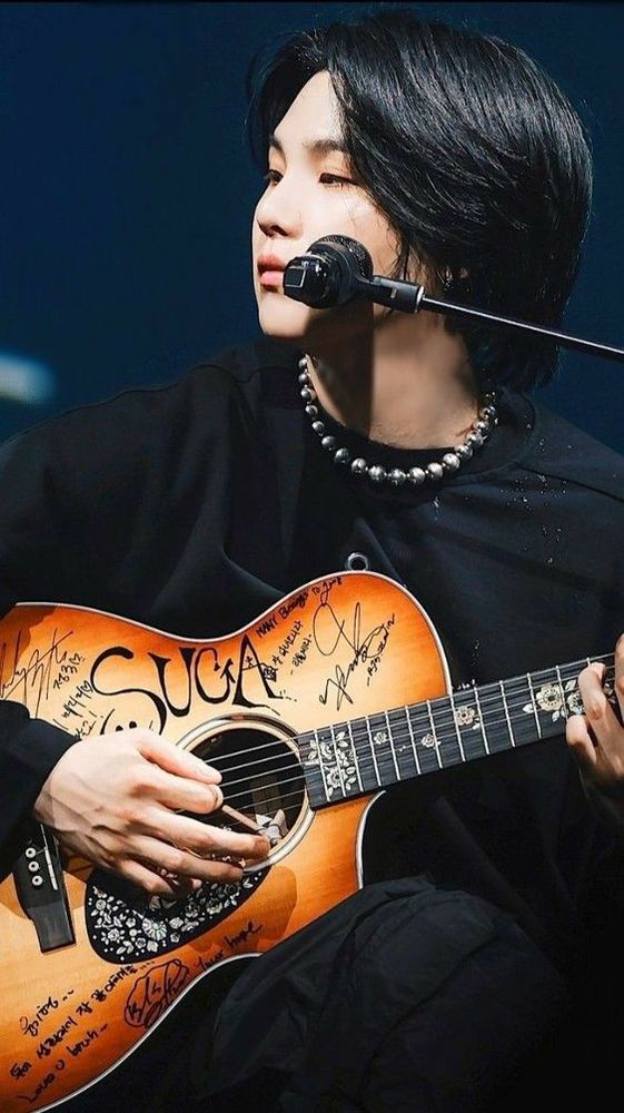 Yoongi está sentado com o violão no colo e olhando para o lado. Ele está vestindo roupas pretas e colar e brincos prateados.

Yoongi is sitting with the guitar on his lap and looking to the side. He is wearing black clothes and a silver necklace and earrings.