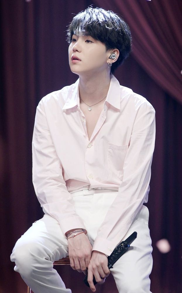 Yoongi está sentado em um banquinho durante uma apresentação e olhando para o lado. Ele está vestindo uma calça branca, camisa social rosa clara e usa um colar e dois pares de brincos de argola prateados. Seu cabelo escuro está combinando com o microfone preto brilhoso.

Yoongi is sitting on a stool during a performance and looking to the side. He is wearing white pants, a light pink dress shirt, and a necklace and two pairs of silver ring earrings. His dark hair matches the shiny black microphone.