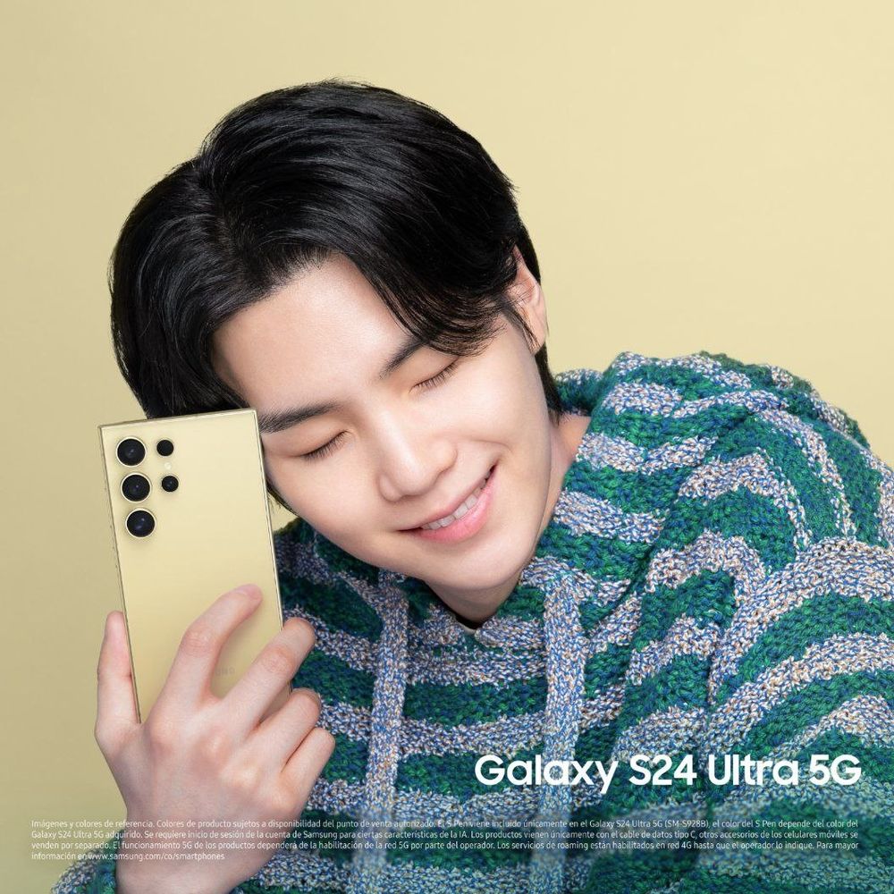Yoongi está segurando um celular enquanto olha para baixo e sorri. Ele usa uma blusa listrada com capuz.

Yoongi is holding a cellphone while looking down and smiling. He is wearing a striped hoodie.