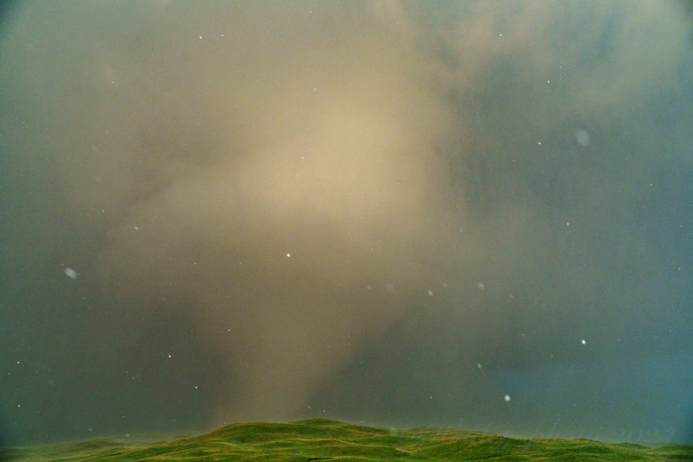 Large hailstones fill the sky in front of a massive tornado partially obscured by the precipitation. The sunset light creates a dramatic scene, and the rolling green sandhills provide a dynamic landscape for such an insane moment.