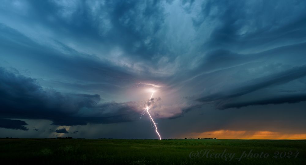 Storm clouds roll above the North Dakota grasslands, a lightning bolt reaches out and hits beyond the horizon. The last orange light of sunset is barely visible behind the blue hued storm.