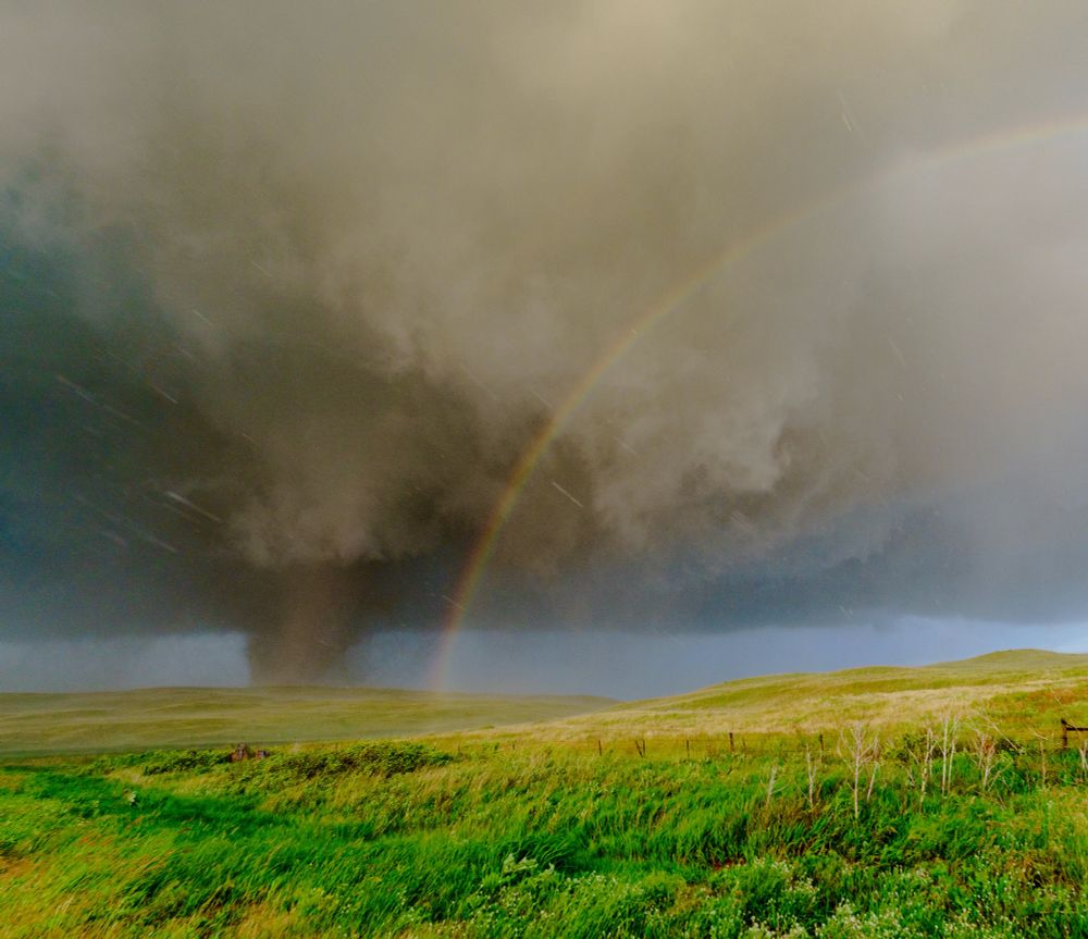 Nebraska. Windblown grasses and brush cover the terrain of the Sandhills as they roll through the foreground. Behind and above the hill to the left a dark brown tornado rages. The tornado is slightly obscured by rain and debris, the clouds above and around it are ragged, gray, and violently rotating. The frame gradually gets brighter from left to right and the left half of a rainbow arcs into the frame and lands just to the right of the tornado.