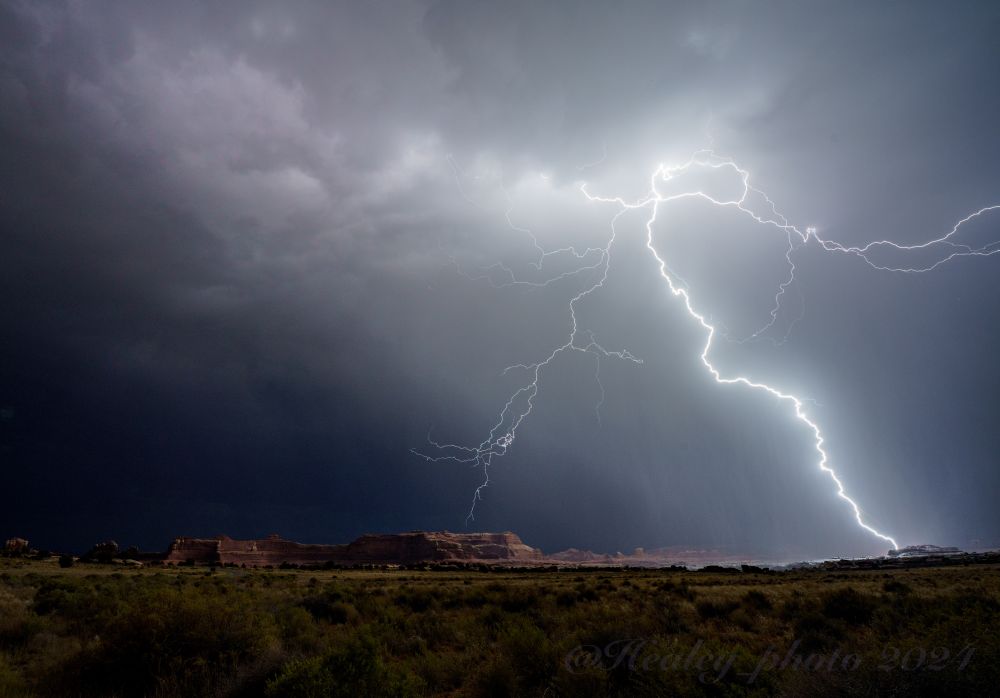 Utah. The monsoon brings a powerful thunderstorm into the Needles district of Canyonlands NP. A huge lightning bolt curves through the air and strikes a rock feature to the right of frame illuminating the landscape below and the rain and clouds above. Lots of red rock terrain features are visible in the distance, steppes and buttes, and then more obscured by rain. In the foreground the shadows of all the desert grasses and brush are cast under the lightning.