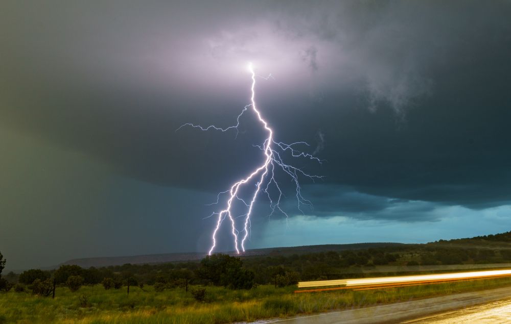 New Mexico. A powerful bolt of lightning strikes the distant hillside with 3 separate branches off the main channel. The clouds and parts of the hill are illuminated by the bolt. The left part of the sky is obscured by rain. In the foreground grassy hills overrun by scrubby brush and plants fill the frame. At bottom right you see the corner of a road and the streaks of headlights as they moved toward the camera while the shutter was open.