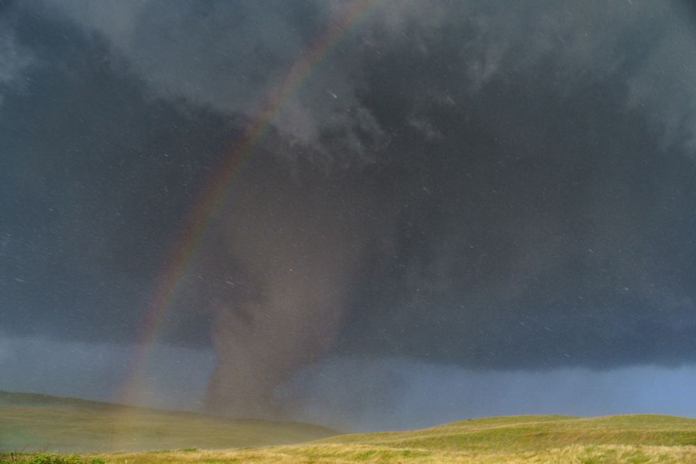 The green hills in the foreground are bright in the sunshine, a ragged dark tornado churns up dirt just beyond the farther hilltop. The sunlight creates a rainbow right in front of the tornado and illuminates individual drops of rain and hailstones flying through the frame.