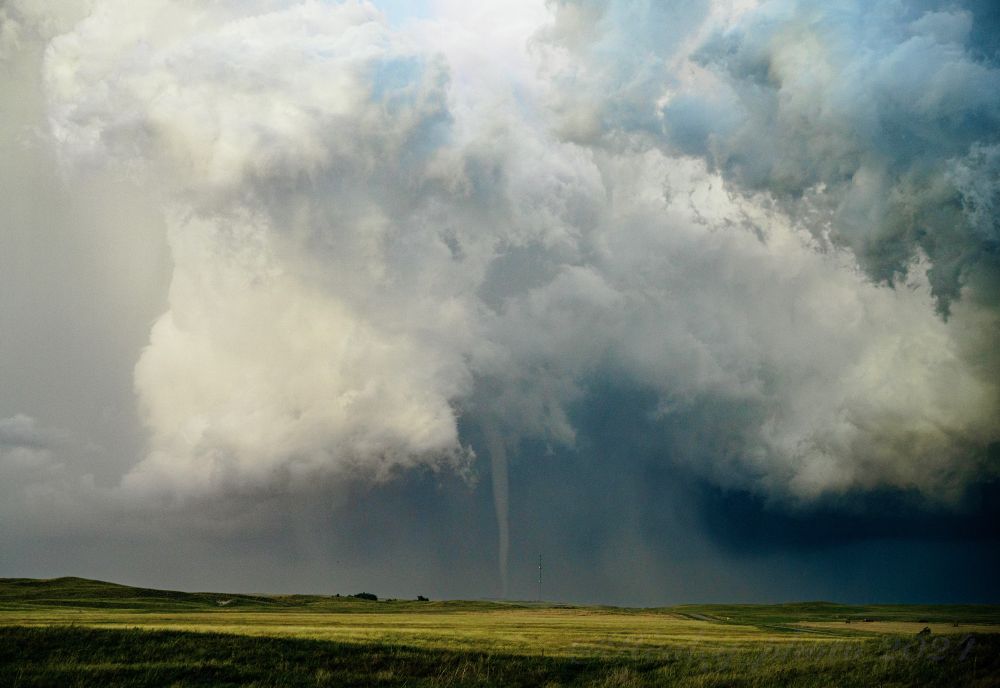 Yellow wheat fields fill a shallow valley between the green sandhills. In the distance a giant white cloud wraps around the skinny shadowed remnants of a significant tornado. A radio tower is almost unnoticeable as it is dwarfed by the tornado behind it.