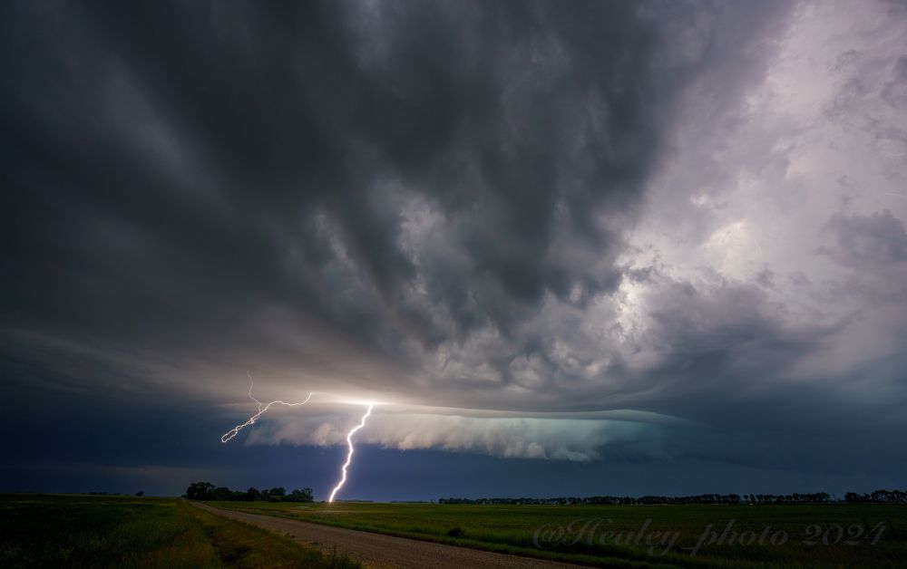 A bright lightning bolt illuminates a low wall cloud as a supercell combs the farmlands of North Dakota. A dirt road ambles towards the storm and over the horizon abutted by bright green fields.
