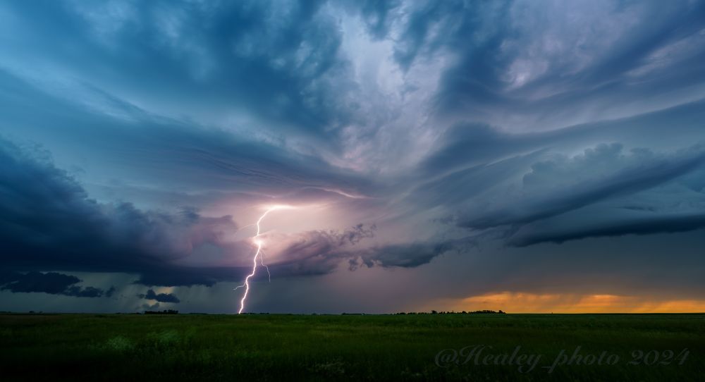 Storm clouds roll above the North Dakota grasslands, a purple tinted lightning bolt reaches out and hits beyond the horizon. The last orange light of sunset is barely visible behind the blue hued storm.