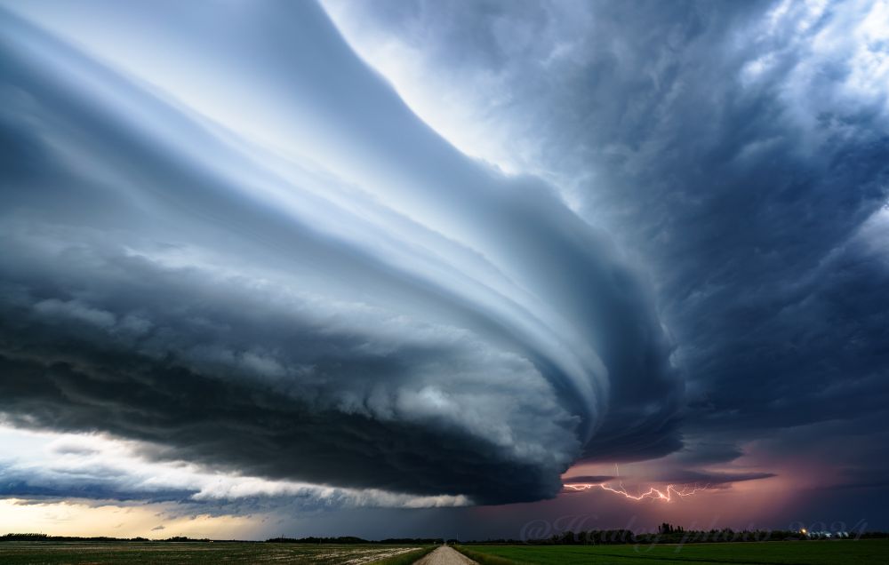 Saskatchewan. The final light of daytime illuminates the back and cloud layers of a sculpted supercell, many of its features are laminar and it has the appearance of stacked plates. The underside of the storm is ragged and dark, with the anvil and distant rain in shadow. Behind the storm infracloud lightning illuminates part of the rain and clouds and hues it a deep purple. A sliver of the foreground is visible showing farm lands, trees a road going straight into the storm and a few farm structures off to the right.