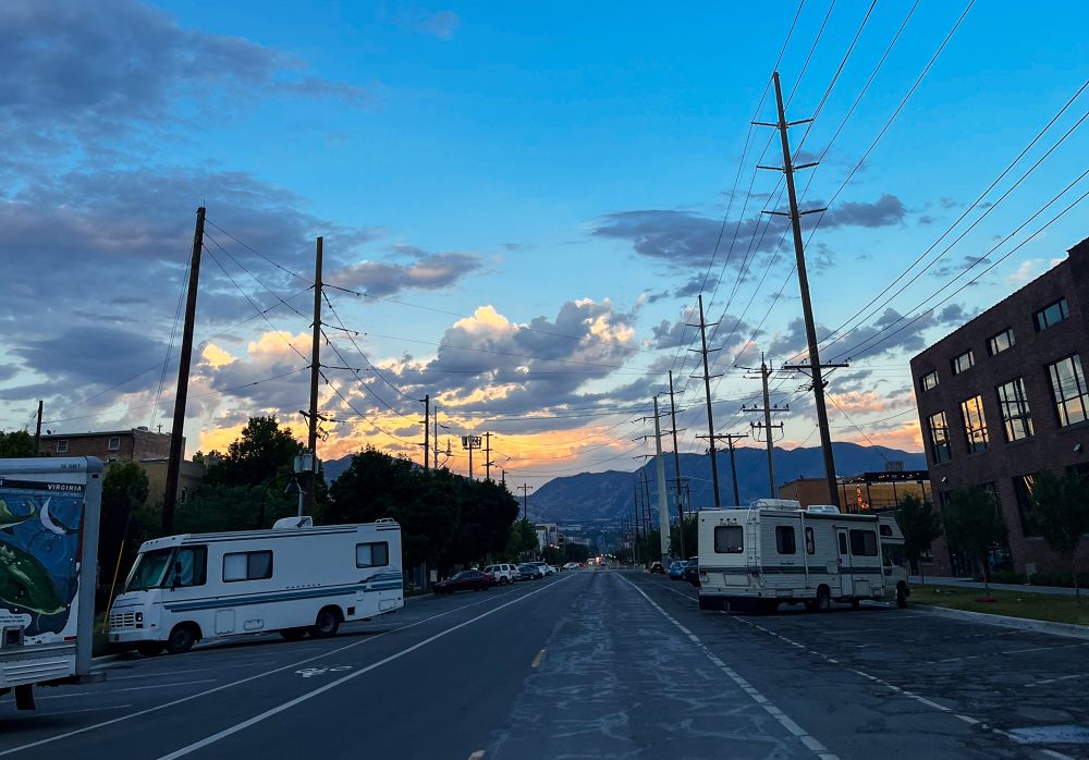 A photo of a road and telephone wires stretching off into Salt Lake City mountains in the distance. Dramatic clouds frame a golden sunset. 