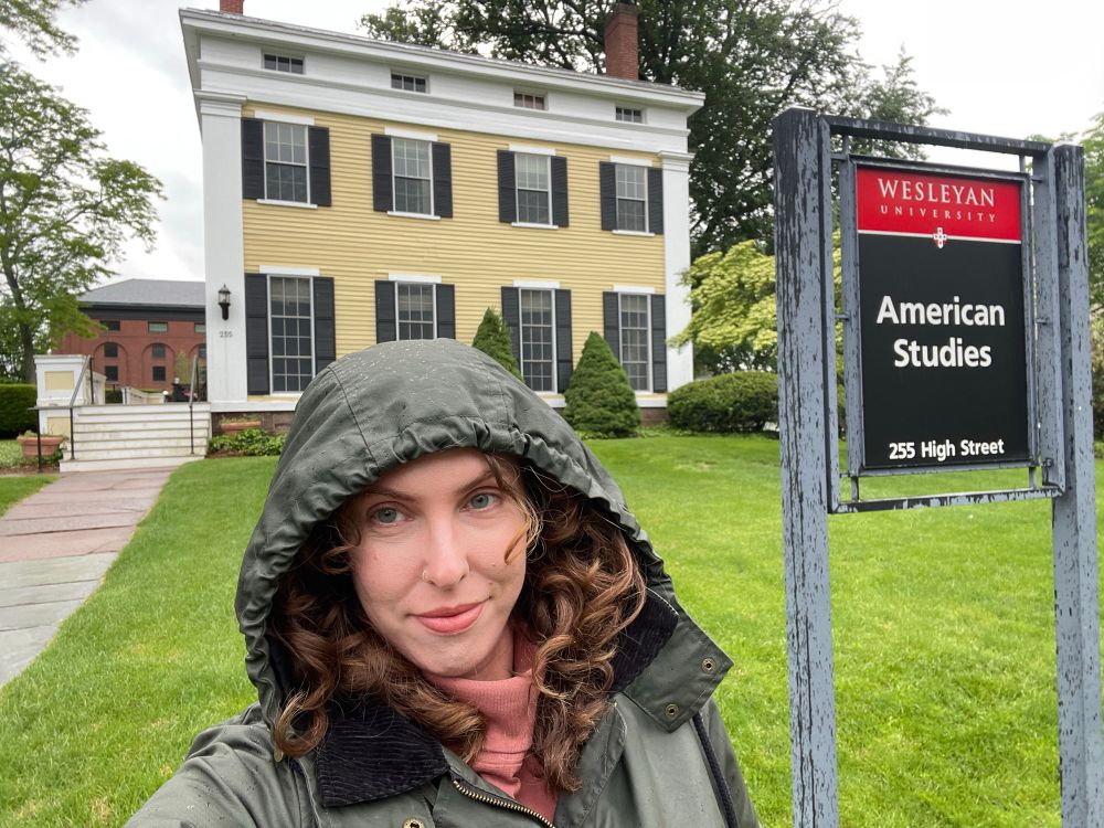 Me, a youngish white woman with curly brown hair in a green hooded raincoat, next to a sign that says “Wesleyan University American Studies,” in front of a yellow and white two-story Adam style house on a bright green lawn. 