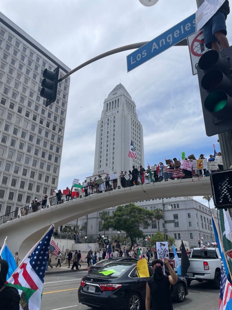 Crowd of protesters on a pedestrian bridge above Los Angeles Street with LA City Hall visible in background. 