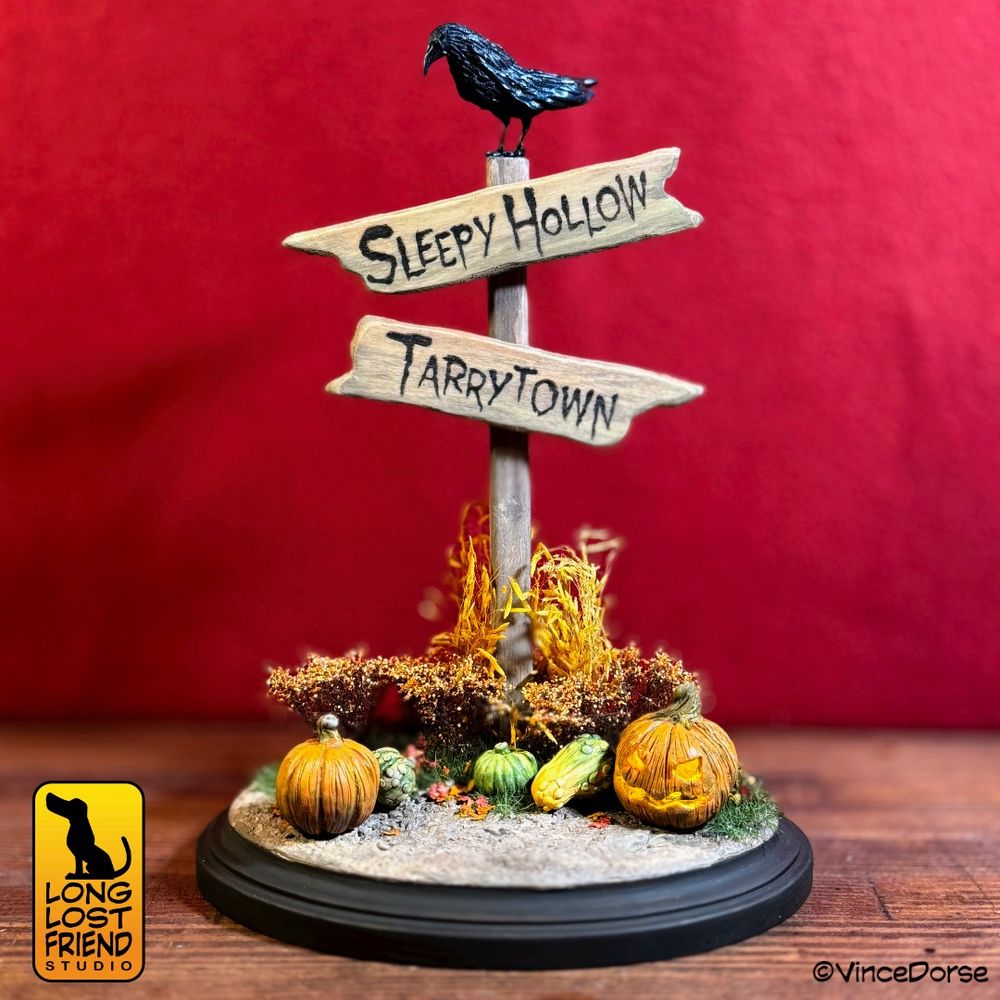 Finished piece of a Sleepy Hollow sign diorama. There is a black bird sitting on top of a sign post. The sign posts has a sign of Sleepy Hollow pointed one way and Tarry Town pointed the other way. Surrounding the bottom of the sign post are various plants and fall foliage. There is a dirt path with horseshoe prints in the path. There are pumpkins, gourds, and a jack o' lantern in front of the plants.
