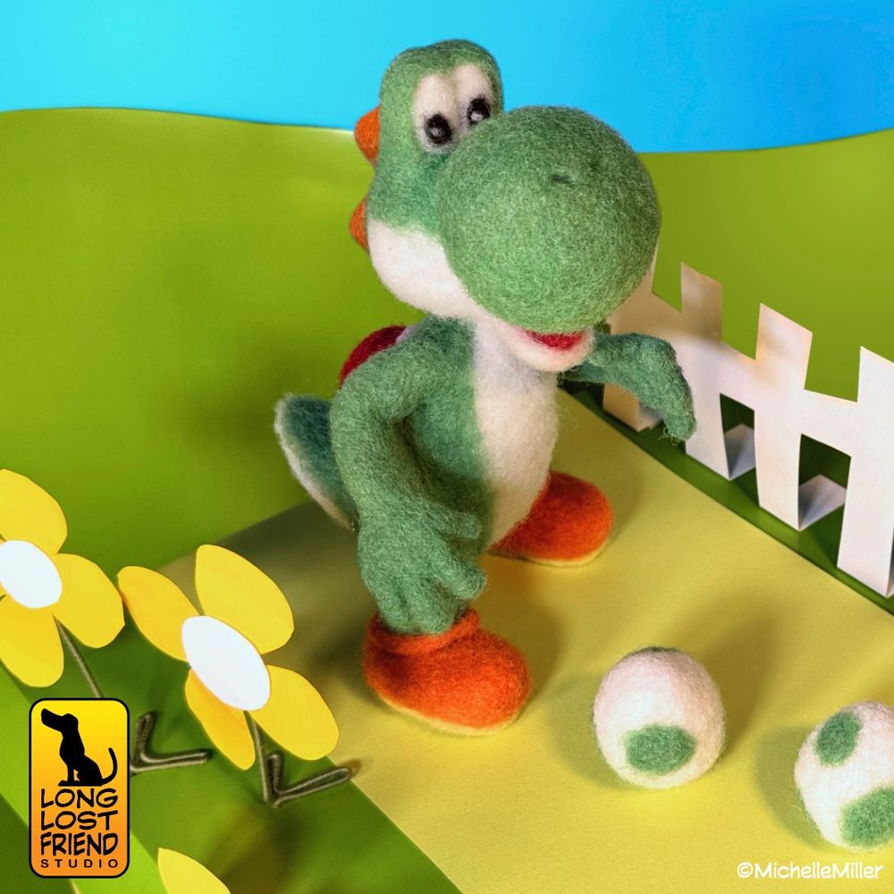 The finished needle felted Yoshi sculpture. Yoshi is in his crafted world with yellow paper flowers on one side of him and a white paper fence is on the other.  He is standing on a yellow paper "road". Yoshi is needle felted by Michelle Miller.