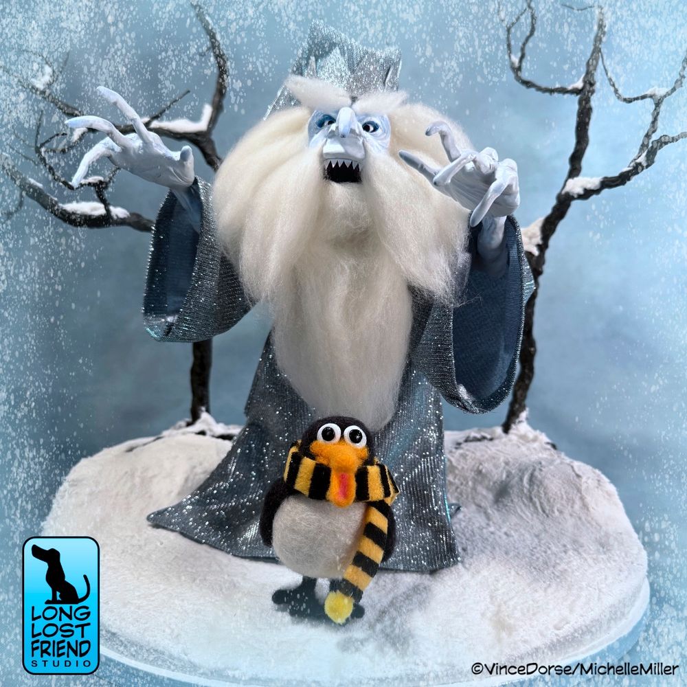 Our finished Winter Warlock art doll and diorama. Winter Warlock is standing in front of two bare, snow-covered trees. Standing in front of Winter Warlock is a needle felted penguin wearing a black and yellow striped scarf --  our version of Topper,  from the Rankin/Bass classic, Santa Claus Is Comin' to Town.