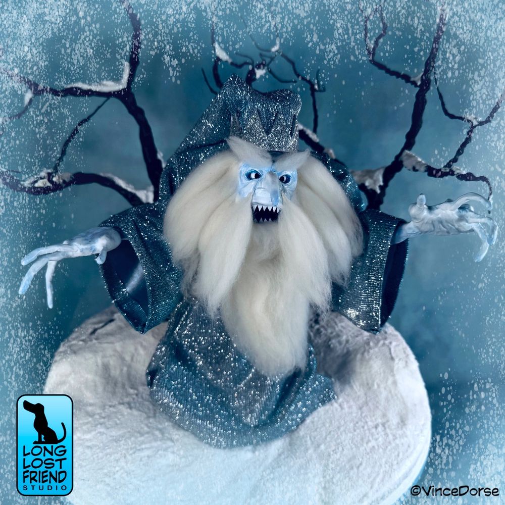 Our finished Winter Warlock Diorama. Winter Warlock art doll is made of clay with long white hair and beard, and a glittery robe and long wizard's hat. He is standing on a snowy base made of foam and has two bare, snow-covered trees behind him made from wire and glue. 