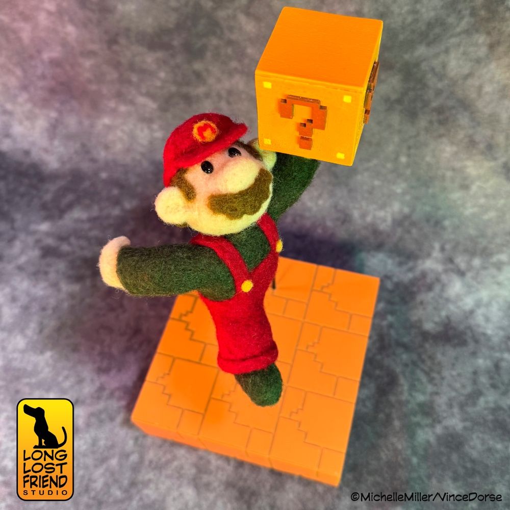 Finished Super Mario diorama - The Plumber. A needle-felted Mario, sporting the colors of his 1985 design, is suspended above a dark orange cube of laser cut terrain, similar to the ground he runs over in the video game. Mario's left hand is raised in a fist and is punching a wooden mystery cube adorned with question marks, as it is in the video game.