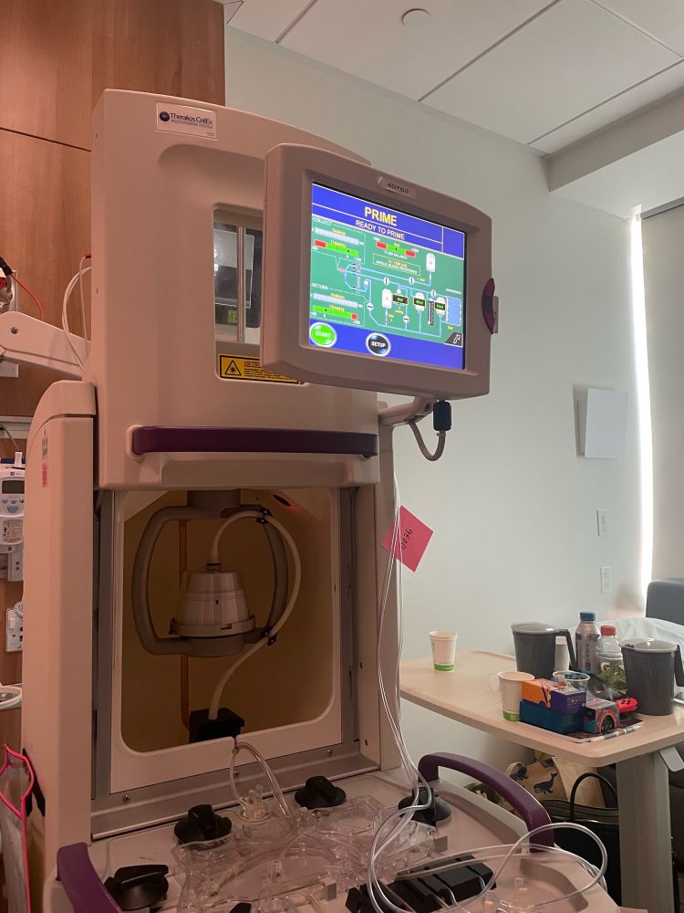 A medical device with a touchscreen displaying “PRIME” and system diagrams, tubes, and components. Background shows a clinical room setup.