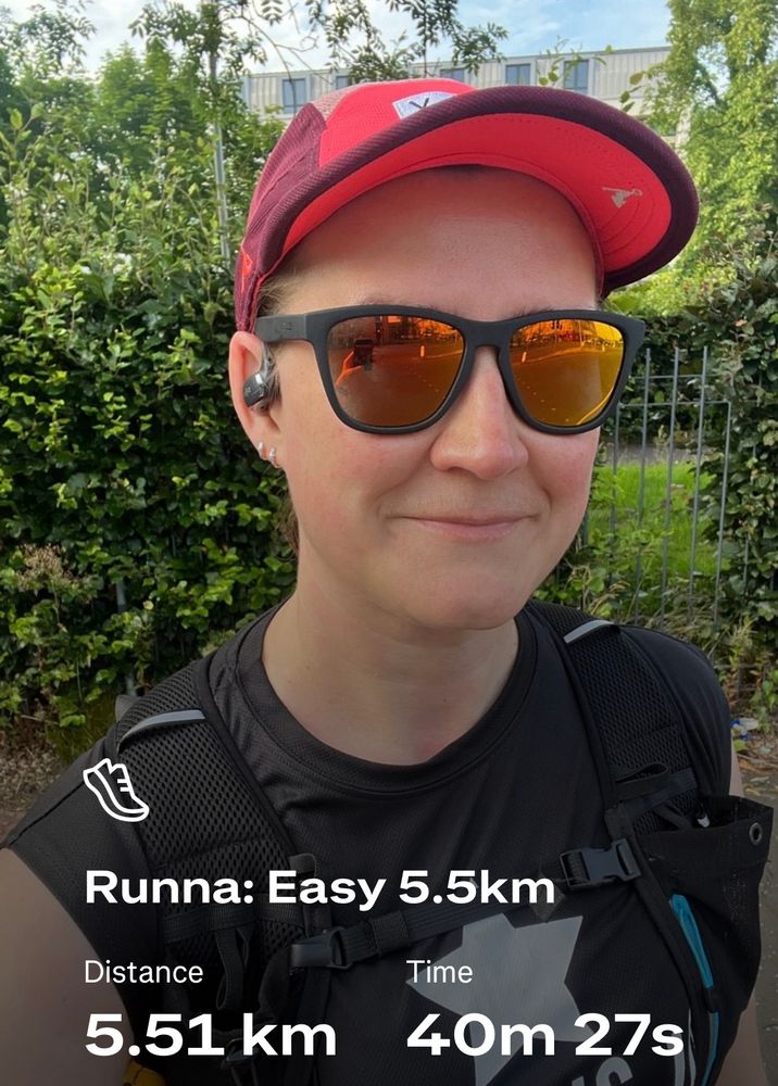 Me, a white woman in my 40’s, taking a post-run selfie outside on a sunny day. I’m wearing a pink baseball cap, black sunglasses, and a black running top and rucksack. I’m smiling at the camera.
