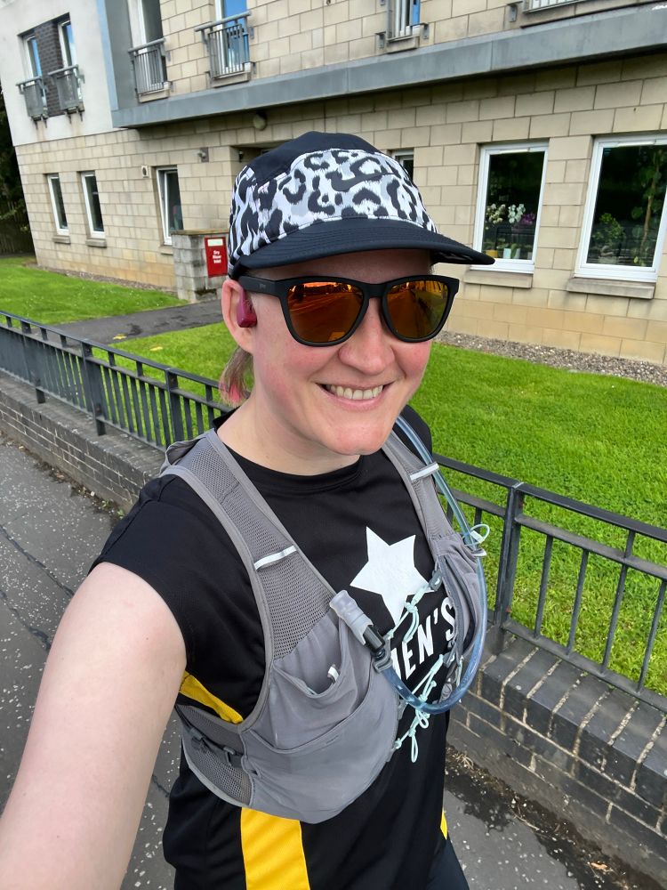 Me, a white woman in my 40’s, taking a selfie outside after a run. I’m wearing a black baseball cap, black sunnies, black tee and shorts, and a grey hydration pack. I’m smiling at the camera and look a bit pink in the face.