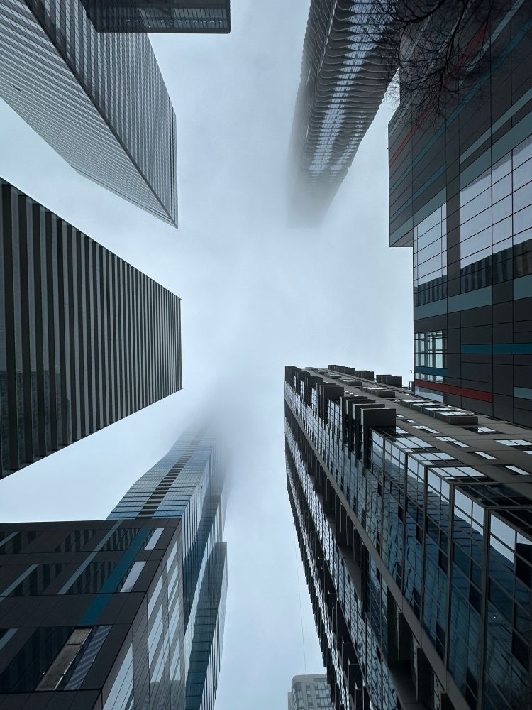 View from base of 7 intricate skyscrapers in close proximity. Fog envelopes the top of the buildings. 