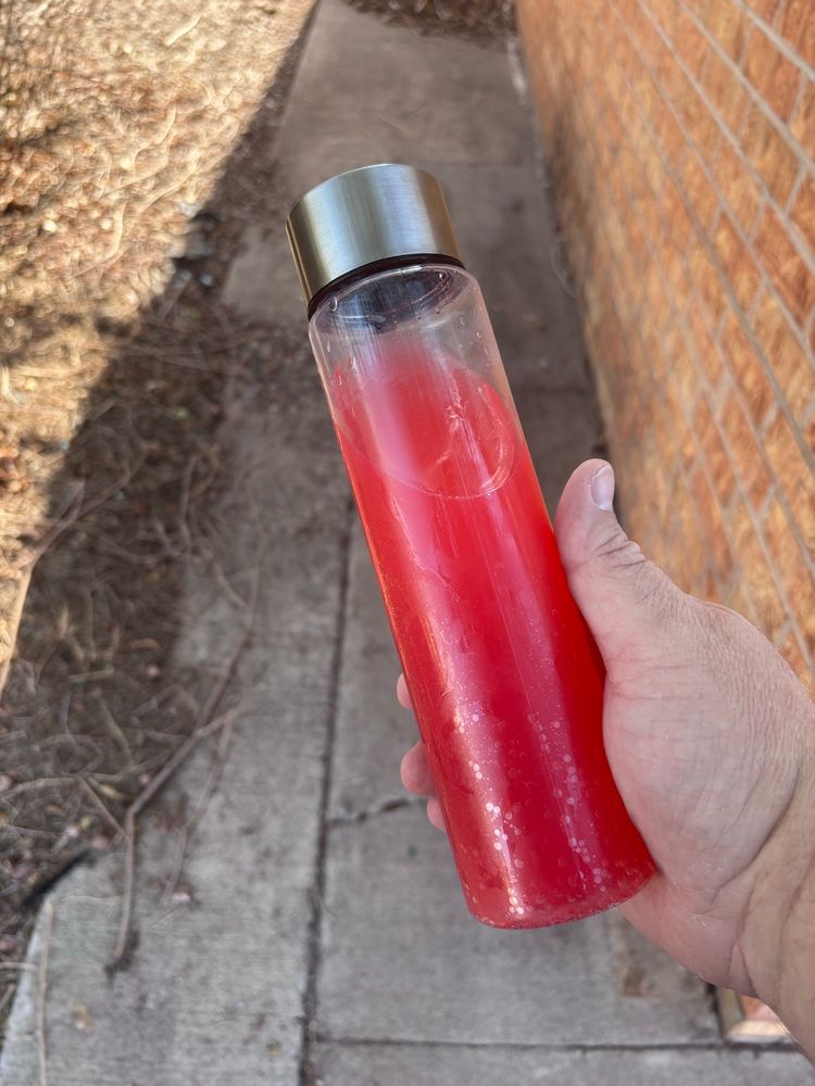 A man's hand made to look fat by the position and angle grips a cylindrical water bottle with no taper, body clear with a chromed cylindrical cap of the same diameter, filled with a deep red liquid. It resembles the canister of future drug Nuke from Robocop 2