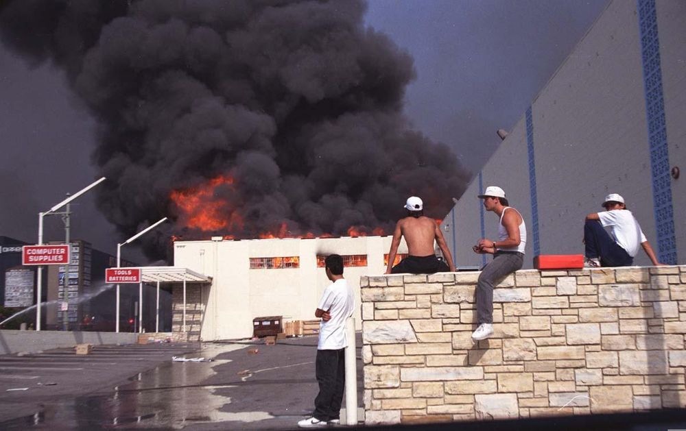 A photo from the Los Angeles riots of 1992. In the foreground are 4 persons, along a rock slate wall. In the background a parking lot and a burning store, pluming with black smoke & flames.