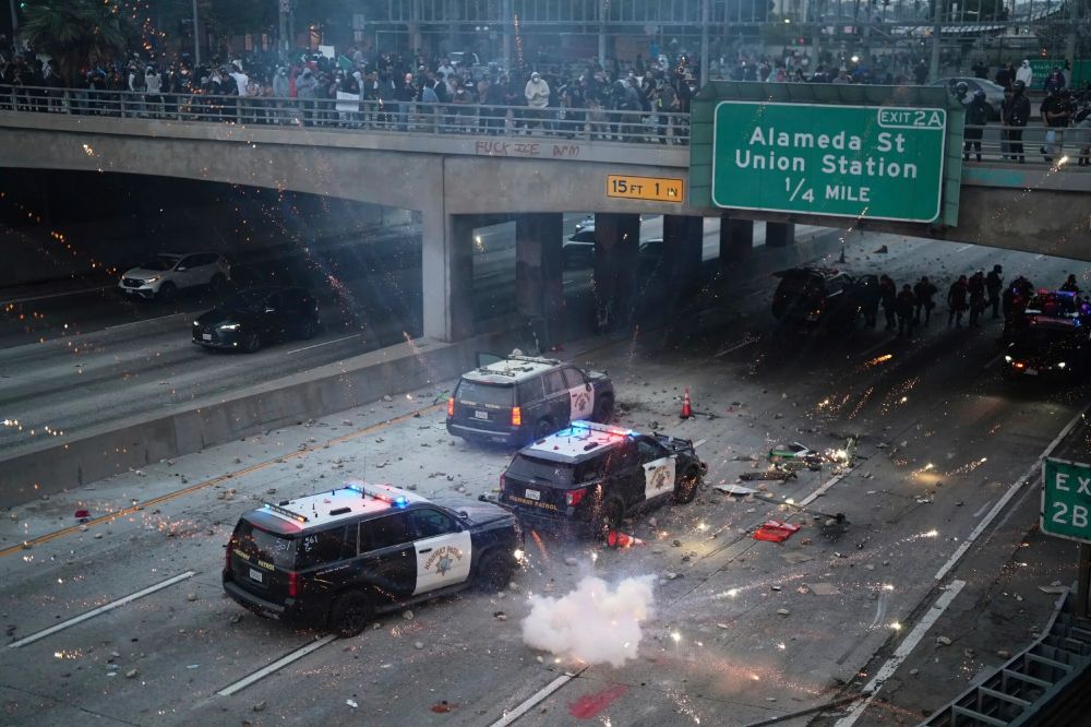 A photograph of the 101 Freeway running through Downtown Los Angeles. Protestors view from a bridge CBP SUVs pelted by rocks, e-scooters & fireworks.